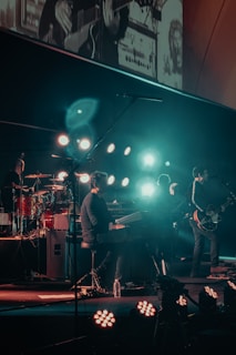 A band performing on stage with various musical instruments, including a keyboard, drum set, and electric guitar. The stage is illuminated by multiple bright lights, creating a vibrant atmosphere. Band members are engaged in playing their instruments, contributing to a dynamic live music setting.