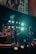 A band performing on stage with various musical instruments, including a keyboard, drum set, and electric guitar. The stage is illuminated by multiple bright lights, creating a vibrant atmosphere. Band members are engaged in playing their instruments, contributing to a dynamic live music setting.