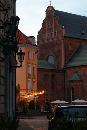 A historic building with a tall brick facade and detailed architecture stands beside a narrow street. Below, a cozy outdoor caf&eacute; is adorned with warm string lights and surrounded by colorful potted plants. Several people are seen walking near the caf&eacute;, and a car is partially visible in the foreground.