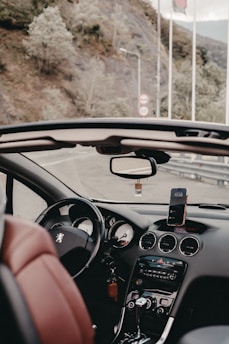 The interior of a convertible car, with a clear view of the steering wheel, dashboard, and controls. A smartphone is mounted on the dashboard, displaying the time. The seats are upholstered in red leather, and the road outside is visible through the open roof, lined with rocky hills and trees. Flags and road signs are observed in the background.