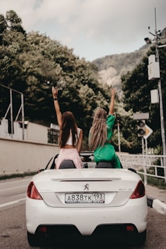 Two people sitting on the back of a white convertible car with their hands raised, as they drive along a road lined with trees. The car's license plate is visible, and the road is bordered by a stone wall and a metal railing.