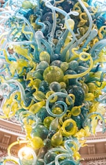 A vibrant, intricate glass sculpture composed of twisting and swirling glass forms in shades of blue, green, and yellow, resembling organic shapes or sea creatures. The sculpture extends upwards, creating a dynamic and lively impression, with each glass piece reflecting light and casting subtle reflections.