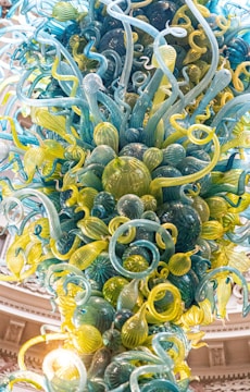 A vibrant, intricate glass sculpture composed of twisting and swirling glass forms in shades of blue, green, and yellow, resembling organic shapes or sea creatures. The sculpture extends upwards, creating a dynamic and lively impression, with each glass piece reflecting light and casting subtle reflections.