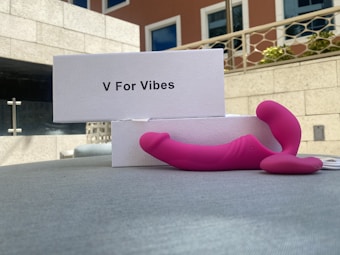 A pink device with a curved design is placed on a surface next to a white box labeled 'V For Vibes'. The background consists of a building with windows and a balcony with plants.