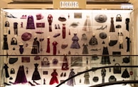 A display case filled with various clothing and accessories, predominantly featuring designs from Dior. The items include dresses, coats, handbags, hats, shoes, and other fashion pieces in shades of purple, red, and black. The display is well-lit, highlighting the elegance and variety of the items showcased.