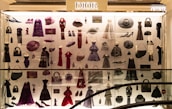 A display case filled with various clothing and accessories, predominantly featuring designs from Dior. The items include dresses, coats, handbags, hats, shoes, and other fashion pieces in shades of purple, red, and black. The display is well-lit, highlighting the elegance and variety of the items showcased.