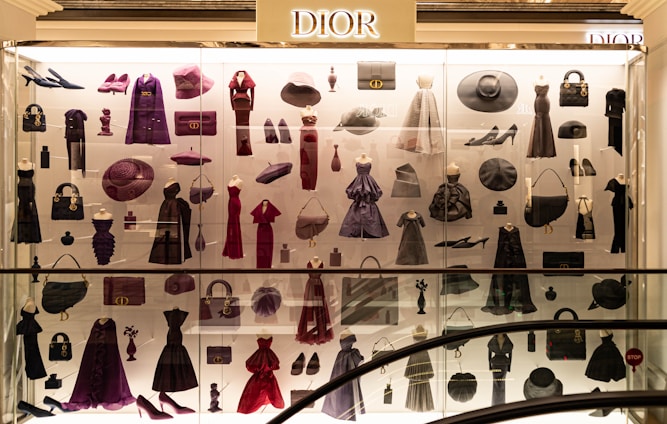 A display case filled with various clothing and accessories, predominantly featuring designs from Dior. The items include dresses, coats, handbags, hats, shoes, and other fashion pieces in shades of purple, red, and black. The display is well-lit, highlighting the elegance and variety of the items showcased.