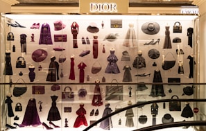 A display case filled with various clothing and accessories, predominantly featuring designs from Dior. The items include dresses, coats, handbags, hats, shoes, and other fashion pieces in shades of purple, red, and black. The display is well-lit, highlighting the elegance and variety of the items showcased.