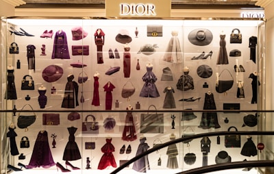 A display case filled with various clothing and accessories, predominantly featuring designs from Dior. The items include dresses, coats, handbags, hats, shoes, and other fashion pieces in shades of purple, red, and black. The display is well-lit, highlighting the elegance and variety of the items showcased.