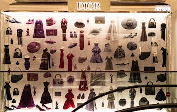 A display case filled with various clothing and accessories, predominantly featuring designs from Dior. The items include dresses, coats, handbags, hats, shoes, and other fashion pieces in shades of purple, red, and black. The display is well-lit, highlighting the elegance and variety of the items showcased.