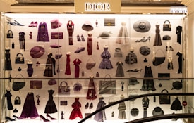 A display case filled with various clothing and accessories, predominantly featuring designs from Dior. The items include dresses, coats, handbags, hats, shoes, and other fashion pieces in shades of purple, red, and black. The display is well-lit, highlighting the elegance and variety of the items showcased.