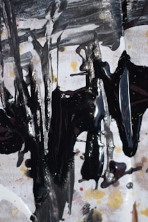 Abstract expressionist artwork featuring bold, dark brushstrokes on a textured background with shades of white, black, and hints of gold and red. The composition creates a dynamic and chaotic pattern, with drips and splatters adding depth and movement.