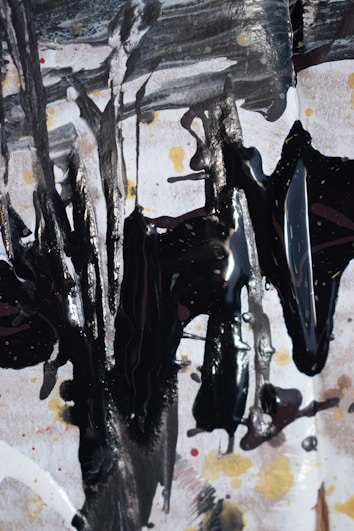 Abstract expressionist artwork featuring bold, dark brushstrokes on a textured background with shades of white, black, and hints of gold and red. The composition creates a dynamic and chaotic pattern, with drips and splatters adding depth and movement.