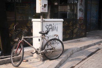 A bicycle is leaning against an electrical box on the sidewalk. The bicycle has a metal frame with noticeable rust and a basket attached to its handlebars. The electrical box has graffiti on it and is situated next to a glass storefront displaying various sea creatures in gold outlines. The pavement is uneven and shows signs of wear.