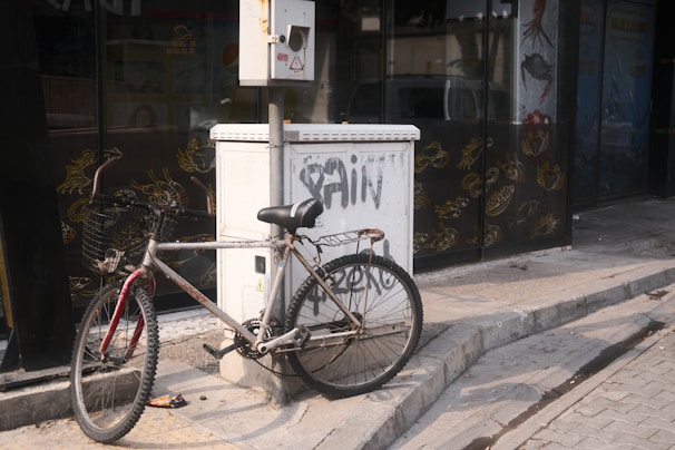 A bicycle is leaning against an electrical box on the sidewalk. The bicycle has a metal frame with noticeable rust and a basket attached to its handlebars. The electrical box has graffiti on it and is situated next to a glass storefront displaying various sea creatures in gold outlines. The pavement is uneven and shows signs of wear.