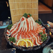 A plate artfully arranged with thinly sliced marbled meat, resembling a mountain shape, is garnished with parsley and cherry tomatoes. Slices of lemon are placed at the base for decoration. A bamboo mat in the background adds an element of texture.