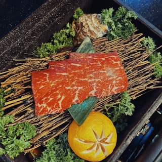 Slices of marbled red meat are artfully arranged on top of a bed of dry twigs garnished with fresh green herbs. A carved orange is placed next to the meat, and a decorative stone is positioned nearby.