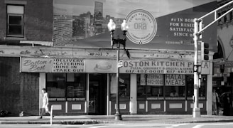 A black and white image of a street view featuring a building with signs for 'Boston's Best' and 'Boston Kitchen Pizza'. There is a person walking on the sidewalk carrying something in their hand. Traffic lights and street lamps are present on the corner. The background includes a large billboard advertisement.