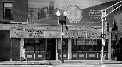 A black and white image of a street view featuring a building with signs for 'Boston's Best' and 'Boston Kitchen Pizza'. There is a person walking on the sidewalk carrying something in their hand. Traffic lights and street lamps are present on the corner. The background includes a large billboard advertisement.