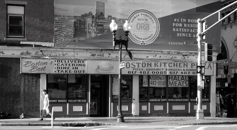 A black and white image of a street view featuring a building with signs for 'Boston's Best' and 'Boston Kitchen Pizza'. There is a person walking on the sidewalk carrying something in their hand. Traffic lights and street lamps are present on the corner. The background includes a large billboard advertisement.