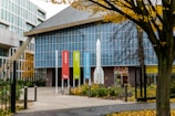 A modern building with a prominent glass facade and geometric design, featuring colorful vertical banners along the entrance path. Trees with autumn foliage surround the area, adding a touch of nature to the urban setting.
