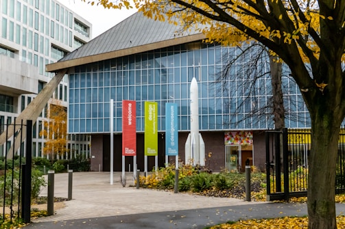 A modern building with a prominent glass facade and geometric design, featuring colorful vertical banners along the entrance path. Trees with autumn foliage surround the area, adding a touch of nature to the urban setting.