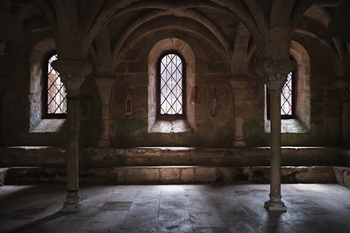 A dimly lit, medieval stone room featuring vaulted ceilings, three tall, narrow windows with intricate leaded glass, stone columns, and bench seating along the walls. The room exudes an ancient and historic atmosphere.