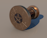A bronze cylindrical object resembling a spool or reel, featuring a circular face with a star-shaped design in the center. The outer edge displays various astrological symbols arranged in a circle. The surface finish appears metallic and polished.