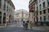 A city street scene features several people walking along a sidewalk. To the right, a London Underground sign is prominently displayed, indicating a subway station entrance. The architecture is characterized by grand, historic buildings with stone exteriors and numerous windows.