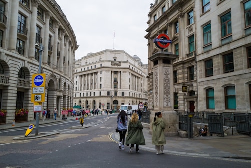 A city street scene features several people walking along a sidewalk. To the right, a London Underground sign is prominently displayed, indicating a subway station entrance. The architecture is characterized by grand, historic buildings with stone exteriors and numerous windows.