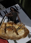 Two luxurious perfume bottles with golden fluid are placed on an ornate gold dish, which also holds a delicate bracelet with gemstones. In the background, a magazine cover is visible, featuring a model in a mesh-patterned dress.