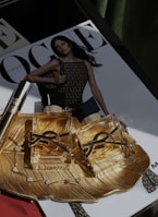 Two luxurious perfume bottles with golden fluid are placed on an ornate gold dish, which also holds a delicate bracelet with gemstones. In the background, a magazine cover is visible, featuring a model in a mesh-patterned dress.