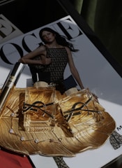 Two luxurious perfume bottles with golden fluid are placed on an ornate gold dish, which also holds a delicate bracelet with gemstones. In the background, a magazine cover is visible, featuring a model in a mesh-patterned dress.