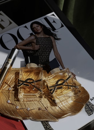 Two luxurious perfume bottles with golden fluid are placed on an ornate gold dish, which also holds a delicate bracelet with gemstones. In the background, a magazine cover is visible, featuring a model in a mesh-patterned dress.