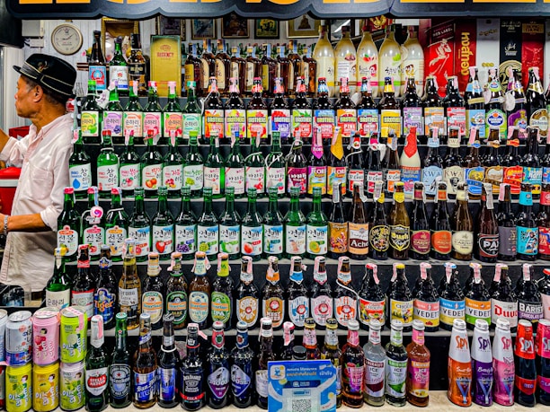A wide variety of alcoholic beverages are displayed on multiple shelves, including beer bottles, cans, and spirits. The colorful array features brands and labels from different regions. To the left, a person in a white shirt and black hat is engaged in organizing or examining the bottles.