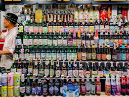 A wide variety of alcoholic beverages are displayed on multiple shelves, including beer bottles, cans, and spirits. The colorful array features brands and labels from different regions. To the left, a person in a white shirt and black hat is engaged in organizing or examining the bottles.