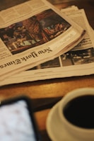 A stack of newspapers, including the New York Times, is placed on a wooden table alongside a cup of coffee. The scene is slightly out of focus, creating a cozy and informal ambiance, with a blurred smartphone visible at the bottom left corner.