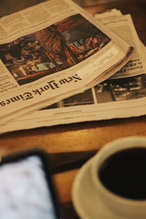 A stack of newspapers, including the New York Times, is placed on a wooden table alongside a cup of coffee. The scene is slightly out of focus, creating a cozy and informal ambiance, with a blurred smartphone visible at the bottom left corner.