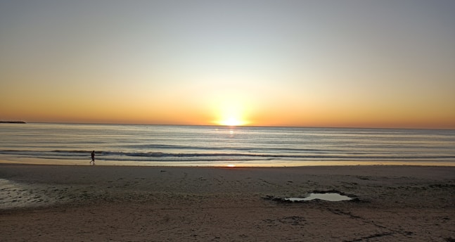 A vibrant sunrise over a tranquil beach with a lone traveler walking along the shore.