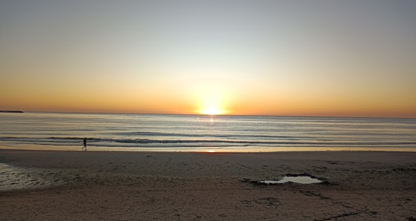 A tranquil beach scene at sunrise with gentle waves lapping against the shore and a lone figure gazing at the horizon.