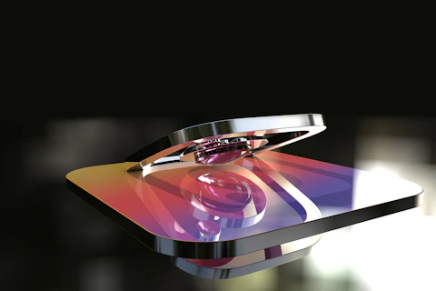 A metallic 3D rendering of a logo with a gradient color scheme featuring hues of purple, orange, and yellow. The logo appears glossy and reflective, with a background that is blurred and dark, highlighting the shiny surface of the object.