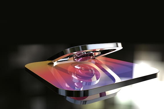 A metallic 3D rendering of a logo with a gradient color scheme featuring hues of purple, orange, and yellow. The logo appears glossy and reflective, with a background that is blurred and dark, highlighting the shiny surface of the object.