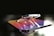 A metallic 3D rendering of a logo with a gradient color scheme featuring hues of purple, orange, and yellow. The logo appears glossy and reflective, with a background that is blurred and dark, highlighting the shiny surface of the object.