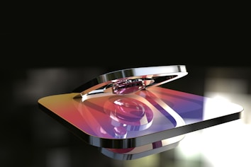 A metallic 3D rendering of a logo with a gradient color scheme featuring hues of purple, orange, and yellow. The logo appears glossy and reflective, with a background that is blurred and dark, highlighting the shiny surface of the object.