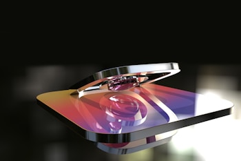 A metallic 3D rendering of a logo with a gradient color scheme featuring hues of purple, orange, and yellow. The logo appears glossy and reflective, with a background that is blurred and dark, highlighting the shiny surface of the object.