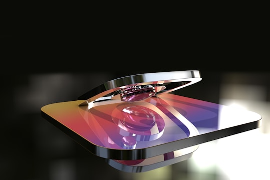 A metallic 3D rendering of a logo with a gradient color scheme featuring hues of purple, orange, and yellow. The logo appears glossy and reflective, with a background that is blurred and dark, highlighting the shiny surface of the object.