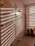 A sophisticated bathroom scene with large hexagonal ceramic tiles on the wall. Three modern gold-colored showerheads are mounted along the tiled wall. Sunlight filters through blinds, creating patterns of light and shadow on the tiles. Two potted snake plants are positioned near a partially visible window with horizontal blinds. The floor is tiled in a lighter stone color, with two white cylindrical containers placed on it.