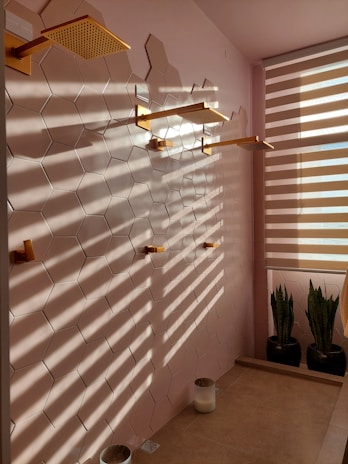 A sophisticated bathroom scene with large hexagonal ceramic tiles on the wall. Three modern gold-colored showerheads are mounted along the tiled wall. Sunlight filters through blinds, creating patterns of light and shadow on the tiles. Two potted snake plants are positioned near a partially visible window with horizontal blinds. The floor is tiled in a lighter stone color, with two white cylindrical containers placed on it.