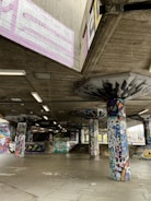 A concrete skatepark covered with vibrant graffiti art on walls, pillars, and ceilings. The space is open with no people present, featuring various shapes and colors in the artwork. Fluorescent lights hang from the ceiling, adding to the urban atmosphere.