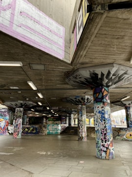 A concrete skatepark covered with vibrant graffiti art on walls, pillars, and ceilings. The space is open with no people present, featuring various shapes and colors in the artwork. Fluorescent lights hang from the ceiling, adding to the urban atmosphere.